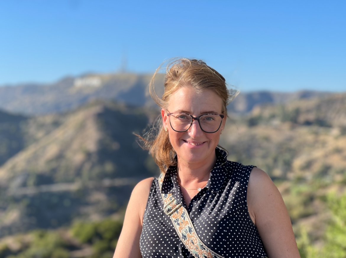 IMG_E53EDAC55E4A-1 A smiling woman with light brown hair and glasses poses outdoors with a mountainous landscape behind her. She is wearing a black sleeveless polka-dot blouse and a patterned crossbody bag strap, with the wind gently blowing her hair. The background features rolling hills and a faintly visible Hollywood sign, suggesting a scenic location in Los Angeles.