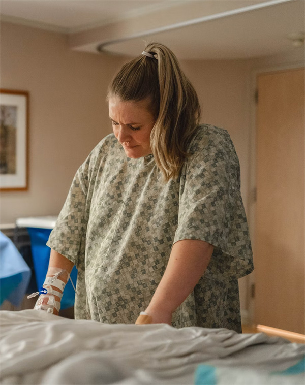 A pregnant woman in a hospital gown leans over a bed during active labor, showing signs of focus and discomfort. An IV is attached to her arm, indicating she is receiving medical care in a hospital setting. The image captures a raw and intimate moment of childbirth preparation.