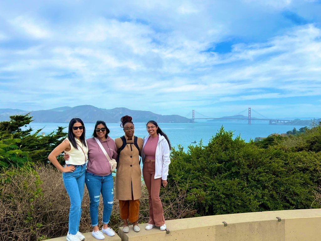 Four women stand together, smiling in front of a scenic view of the Golden Gate Bridge and the San Francisco Bay. They are dressed in casual and stylish outfits, with sunglasses, crossbody bags, and jackets. The sky is partly cloudy, and lush greenery surrounds them as they pose near a beige stone railing.