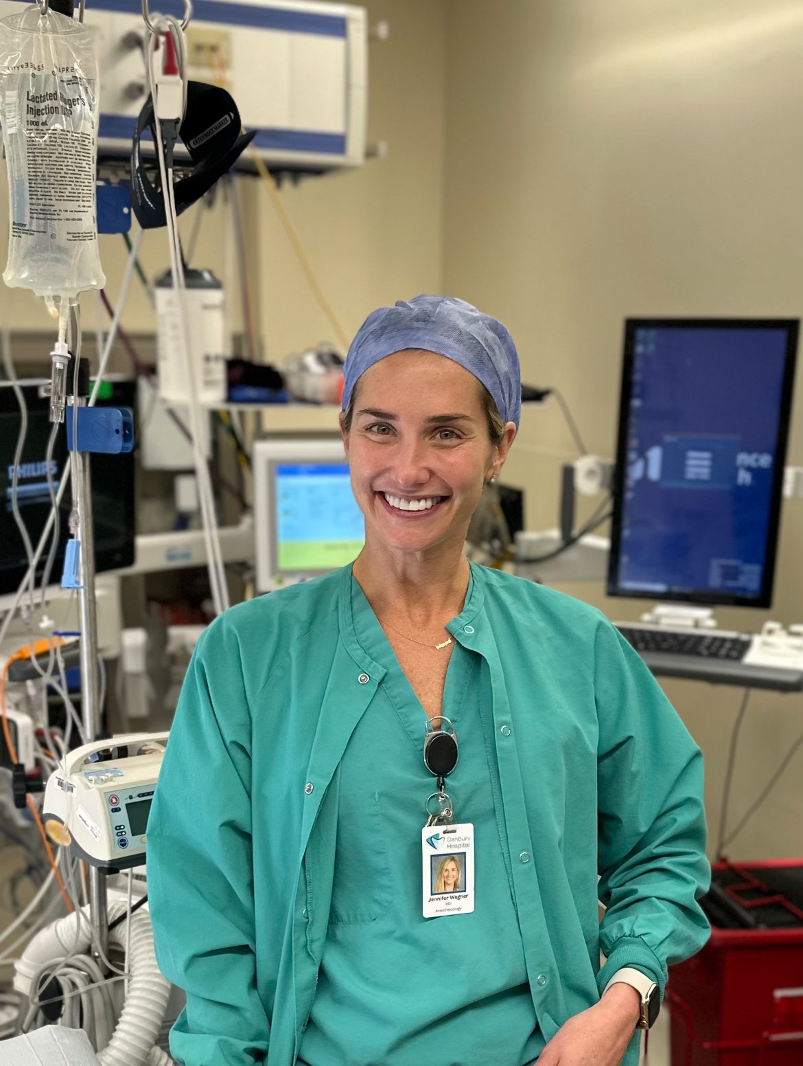 A smiling healthcare professional wearing green surgical scrubs and a blue scrub cap stands in a medical setting. She has a hospital ID badge clipped to her scrubs, with sunglasses hanging from the collar. Behind her, medical monitors, IV bags, and various anesthesia equipment are visible, indicating a hospital or operating room environment.
