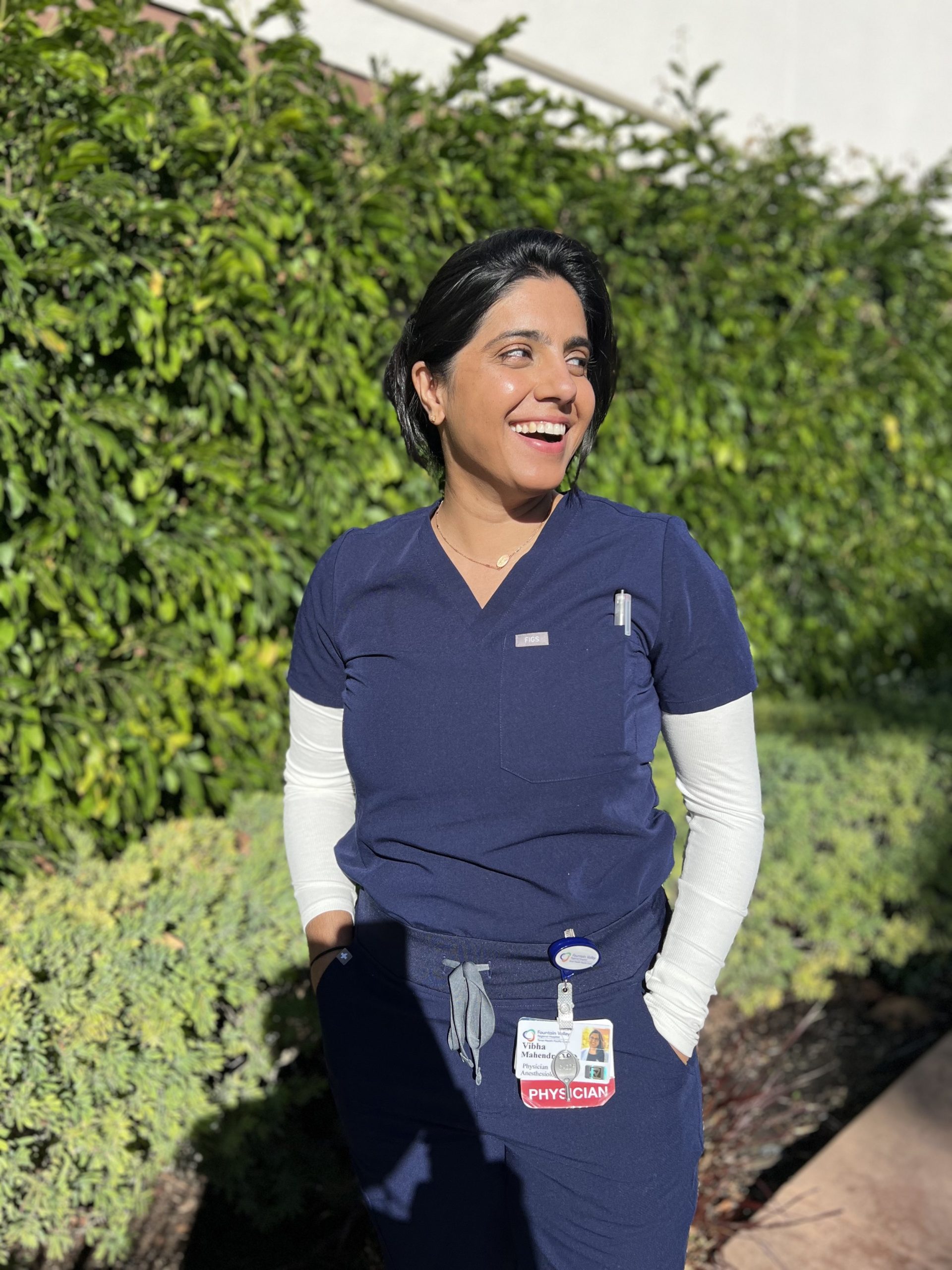 A smiling healthcare professional wearing navy blue scrubs and a white long-sleeve undershirt stands outdoors in bright sunlight. She has short black hair, hands in her pockets, and an ID badge clipped to her scrubs that reads "PHYSICIAN." Lush green foliage and a white building wall are visible in the background.