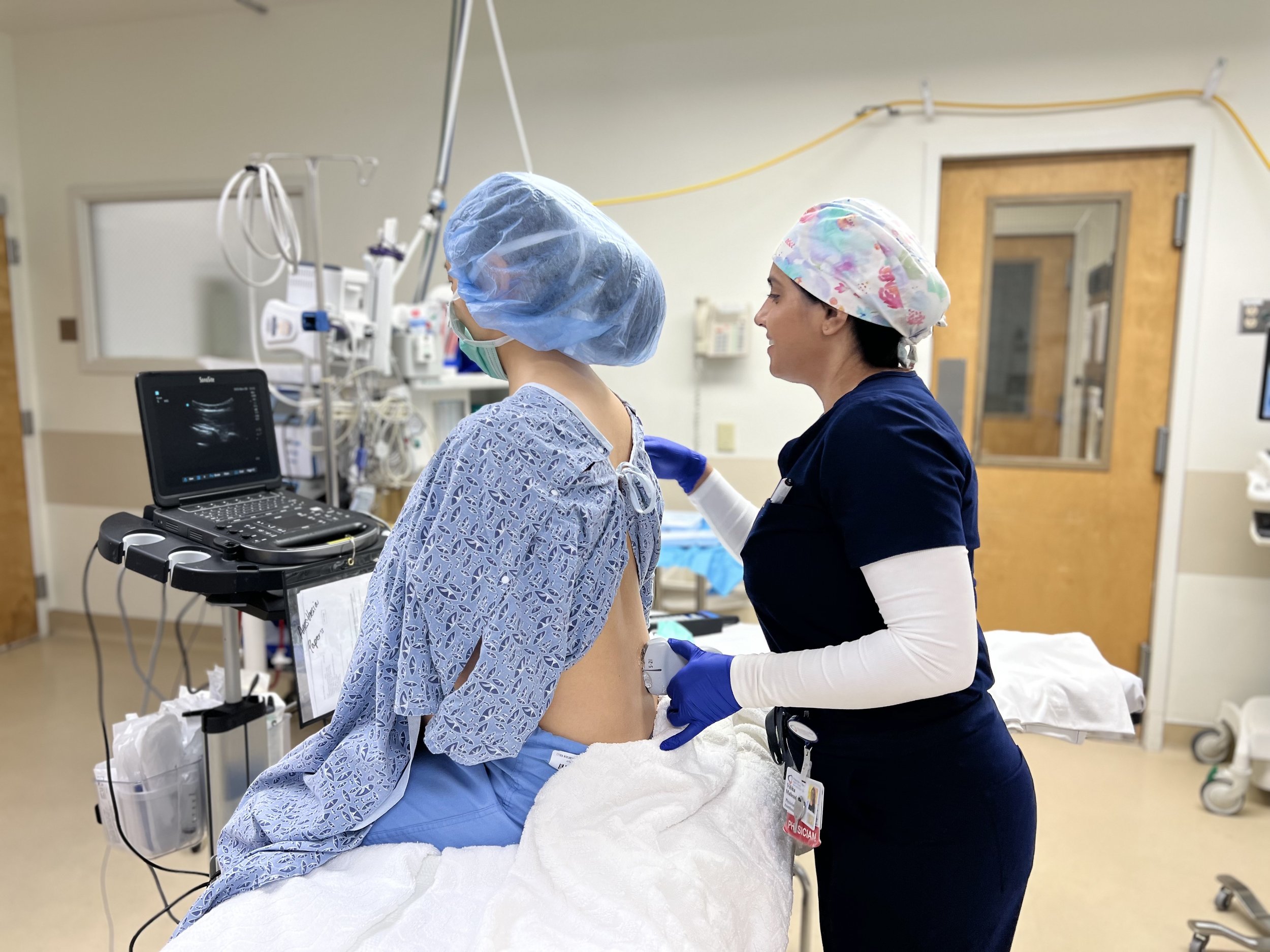 A healthcare professional in navy blue scrubs and a floral surgical cap prepares a patient for a procedure in a hospital setting. The patient, wearing a blue surgical gown and a hairnet, sits on a hospital bed with their back exposed while the professional holds an ultrasound probe near their lower back. A portable ultrasound machine displays an image on the screen, and various medical equipment is visible in the background.