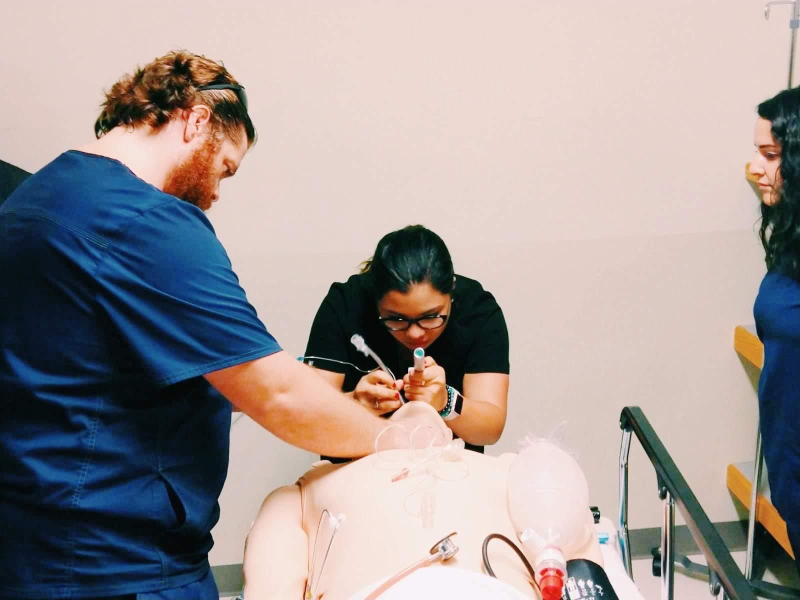 Three healthcare professionals practice an intubation procedure on a medical mannequin in a clinical training setting. One person in black scrubs is carefully inserting a tube into the mannequin’s airway, while another in navy blue scrubs assists. A third person observes from the side. Various medical instruments and airway management tools are visible on the table.