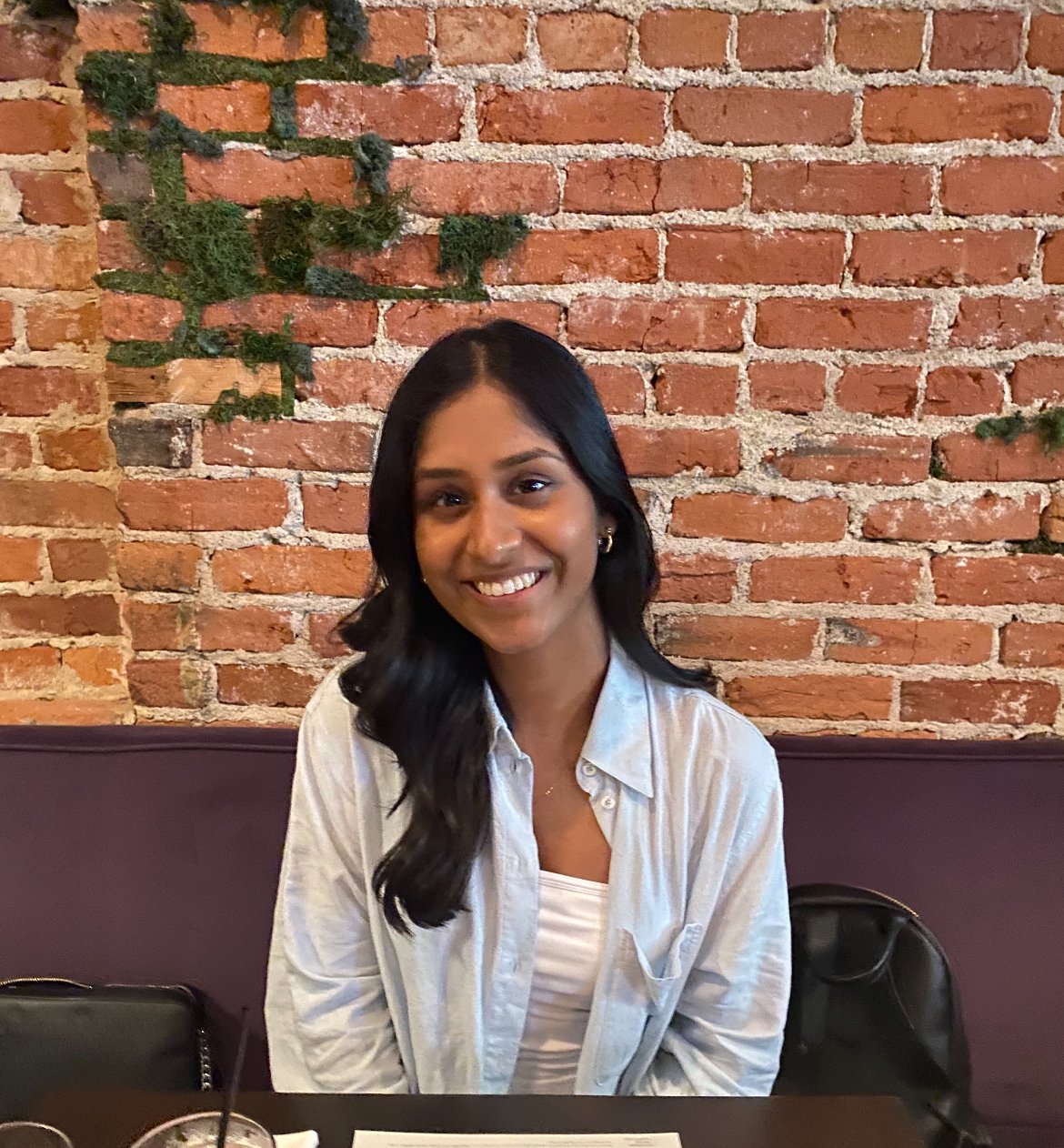 A young woman with long, dark hair and a warm smile sits at a restaurant table, looking at the camera. She is wearing a light blue button-up shirt over a white top, with a brick wall decorated with patches of greenery in the background. A menu, a drink with a straw, and a black purse are visible on the table in front of her.