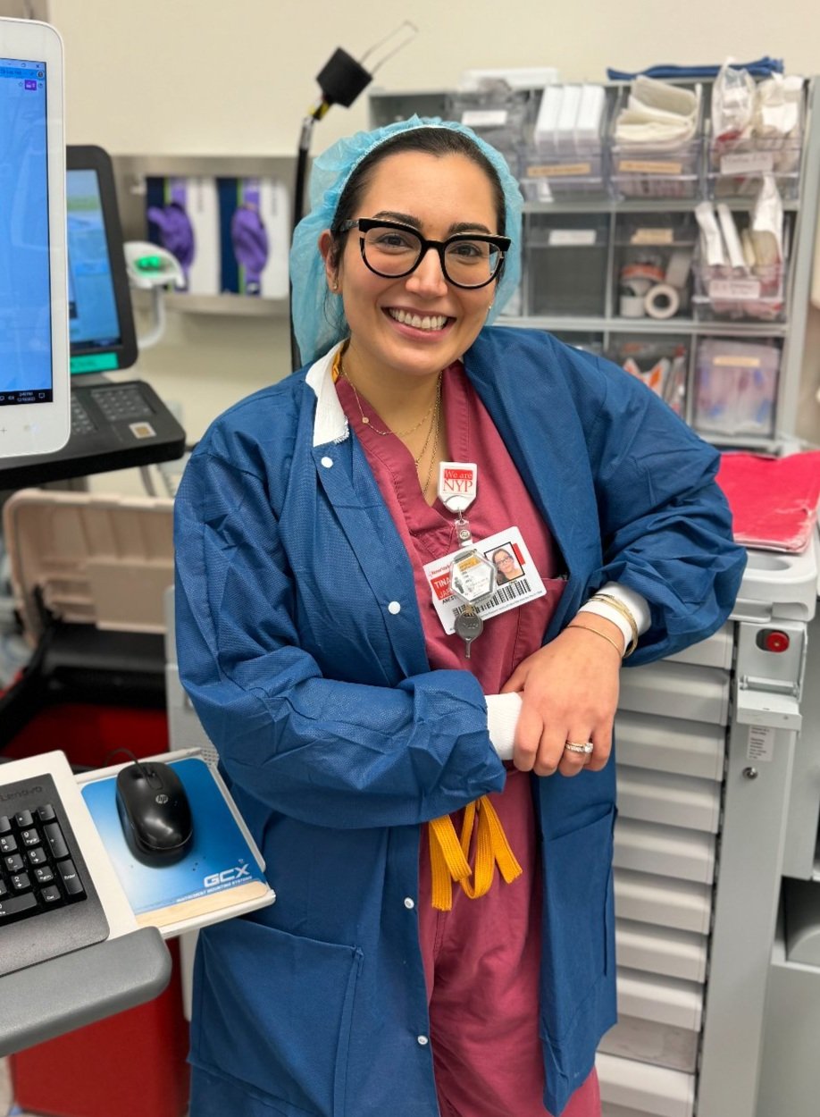 A smiling healthcare professional wearing burgundy scrubs, a blue protective gown, and a hairnet leans on a medical cart in a hospital setting. She has black-framed glasses and an ID badge clipped to her scrubs, displaying her name and affiliation with NewYork-Presbyterian (NYP). Behind her, shelves stocked with medical supplies, a computer monitor, and various hospital equipment are visible.